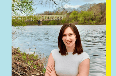 A woman with brown hair stands with arms crossed by a river, with a bridge and trees in the background. The image has a blue and yellow border.