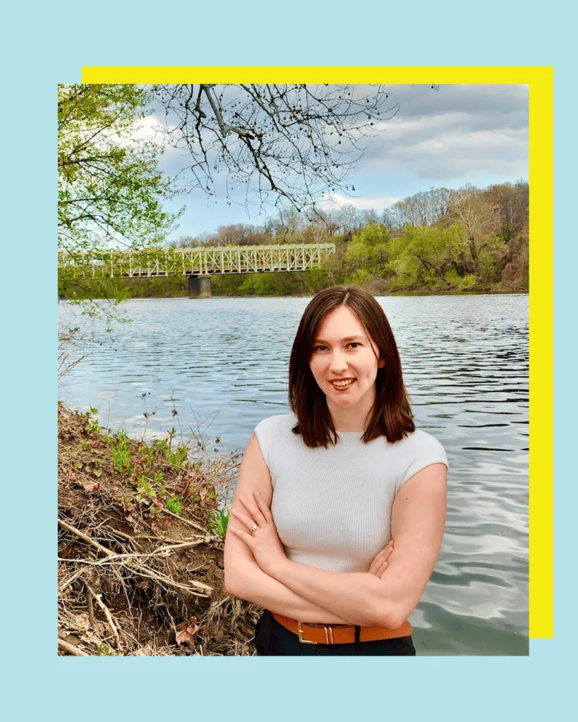 A woman with brown hair stands with arms crossed by a river, with a bridge and trees in the background. The image has a blue and yellow border.