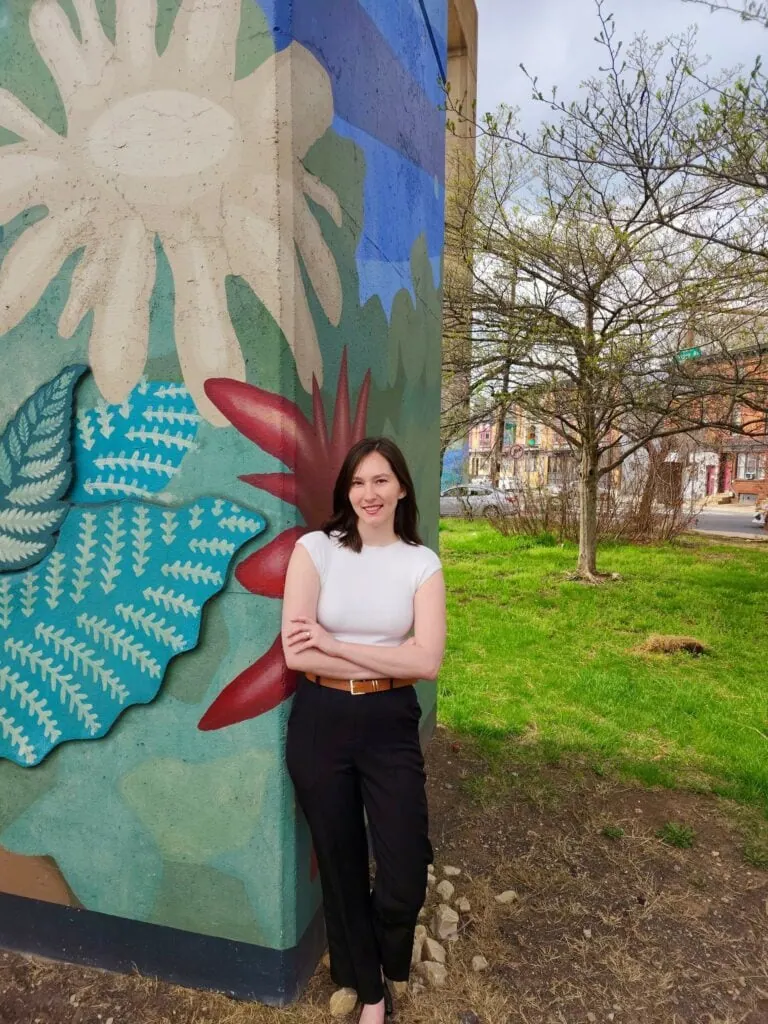 A woman in a white shirt and black pants stands smiling against a colorful mural on a concrete wall, with grass and trees in the background.